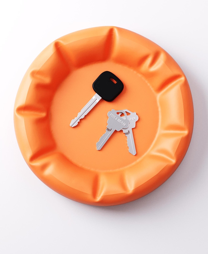 May include: An orange, round catch-all tray with a ridged edge. Two keys rest inside the tray: one with a black head and the other two with silver heads. The keys are likely for a house or car.