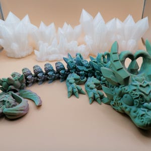 Op de afbeelding: Een gedetailleerde, teal-gekleurde drakenbeeldhouwkunst met ingewikkelde gravures en kristalachtige accenten. De kop en het lichaam van de draak zijn zichtbaar, samen met een apart staartstuk. Op de achtergrond staan ​​verschillende witte, kristalvormige sculpturen.