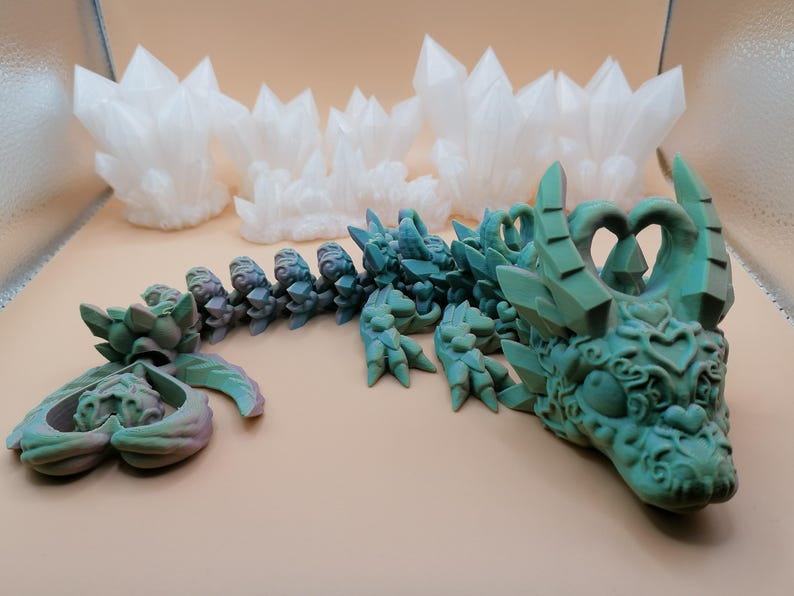 Op de afbeelding: Een gedetailleerde, teal-gekleurde drakenbeeldhouwkunst met ingewikkelde gravures en kristalachtige accenten. De kop en het lichaam van de draak zijn zichtbaar, samen met een apart staartstuk. Op de achtergrond staan ​​verschillende witte, kristalvormige sculpturen.