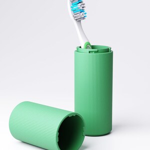 May include: A light green cylindrical toothbrush case with a ribbed texture. One half of the case stands upright containing a white toothbrush with blue and white bristles, while the other half of the case lies horizontally in the foreground.