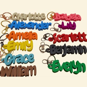 May include: A collection of colorful name keychains in various colors, including beige, blue, orange, yellow, teal, brown, pink, purple, red, black, and green. Each keychain features a different name in a bold, playful font, attached to a metal keyring.