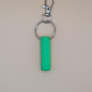 May include: A bright green cylindrical keychain charm attached to a silver metal split ring and a lobster clasp. The charm is a solid, vibrant green color. The background is a solid, neutral beige.