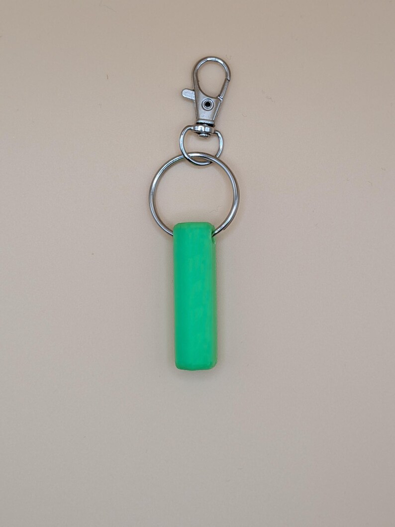 May include: A bright green cylindrical keychain charm attached to a silver metal split ring and a lobster clasp. The charm is a solid, vibrant green color. The background is a solid, neutral beige.