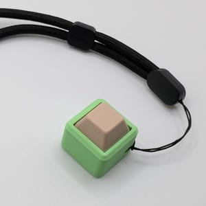 May include: A square, light green device with a tan, pyramid-shaped button. The device is attached to a black lanyard with black plastic adjusters. The background is plain white.