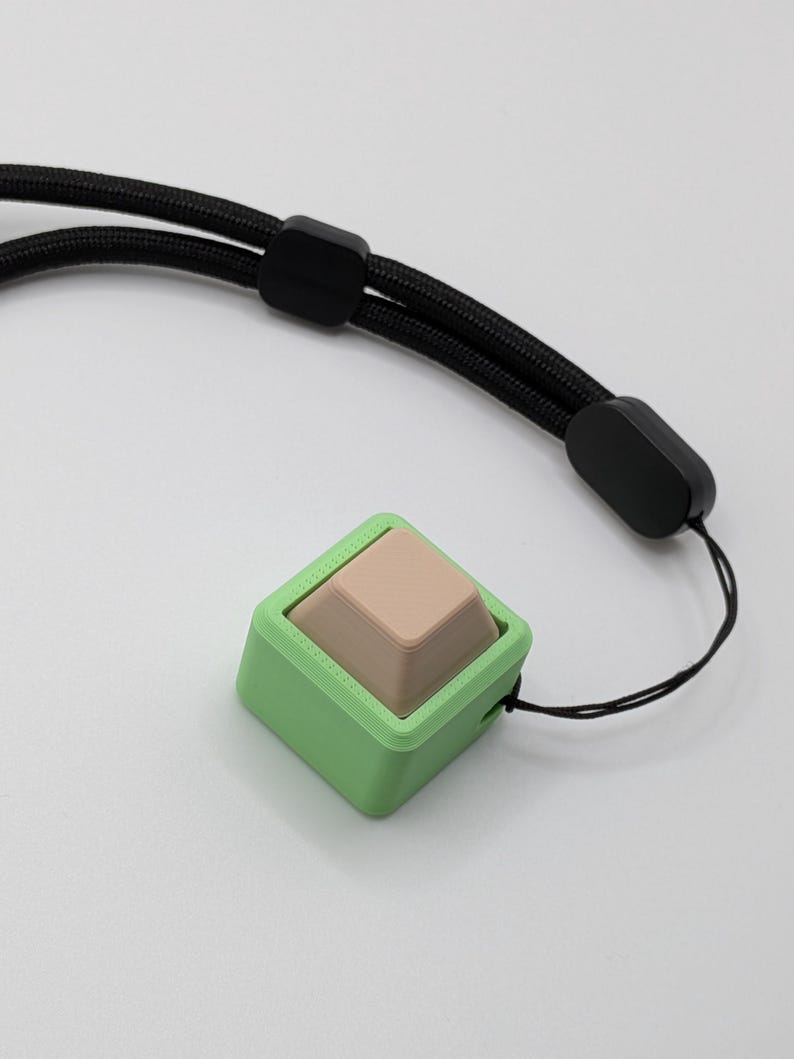 May include: A square, light green device with a tan, pyramid-shaped button. The device is attached to a black lanyard with black plastic adjusters. The background is plain white.