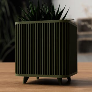 May include: An olive green planter with a square shape and vertical slat design. The planter, supported by four small legs, contains a dark green succulent. The planter is made of wood and is on a wooden table.