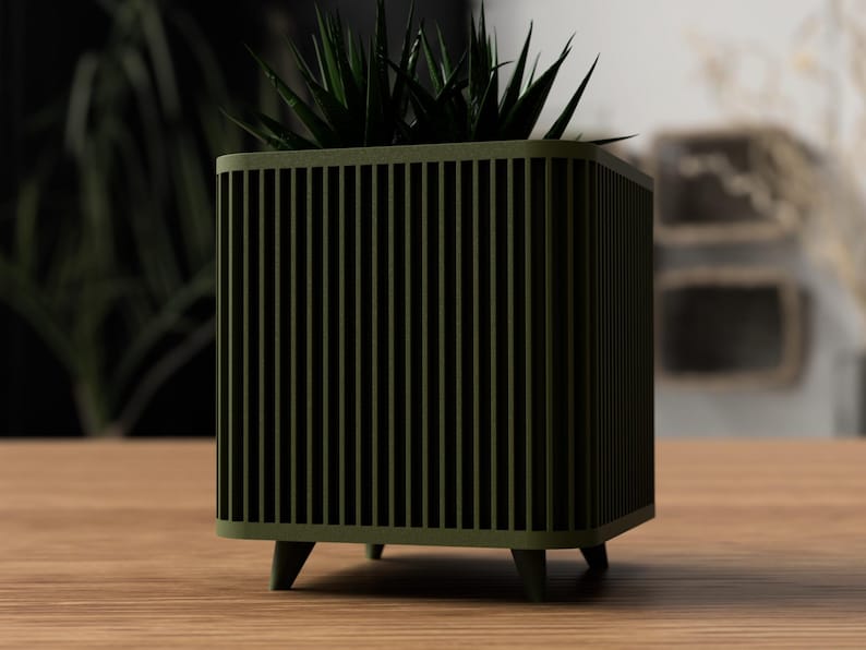 May include: An olive green planter with a square shape and vertical slat design. The planter, supported by four small legs, contains a dark green succulent. The planter is made of wood and is on a wooden table.