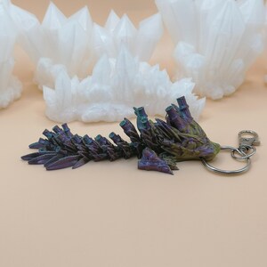 May include: A keychain featuring a detailed, iridescent crystal formation in shades of purple, green, and gold. The keychain has a silver-colored ring and clasp. White crystal clusters are in the background.