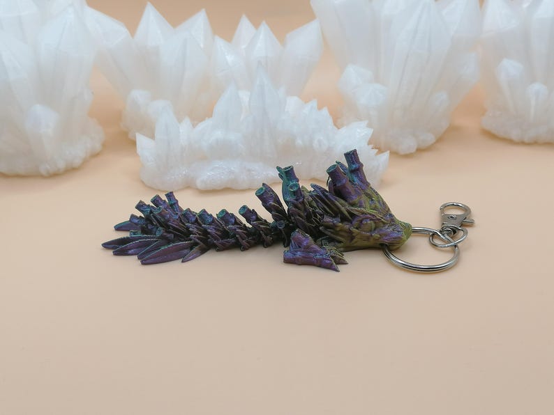 May include: A keychain featuring a detailed, iridescent crystal formation in shades of purple, green, and gold. The keychain has a silver-colored ring and clasp. White crystal clusters are in the background.