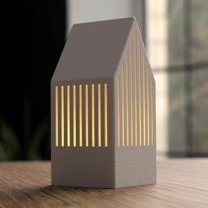 May include: A gray, house-shaped lamp with vertical, illuminated slots. The lamp has a textured base and a pitched roof. The warm light shines through the slots, creating a cozy ambiance. The lamp is on a wooden surface.