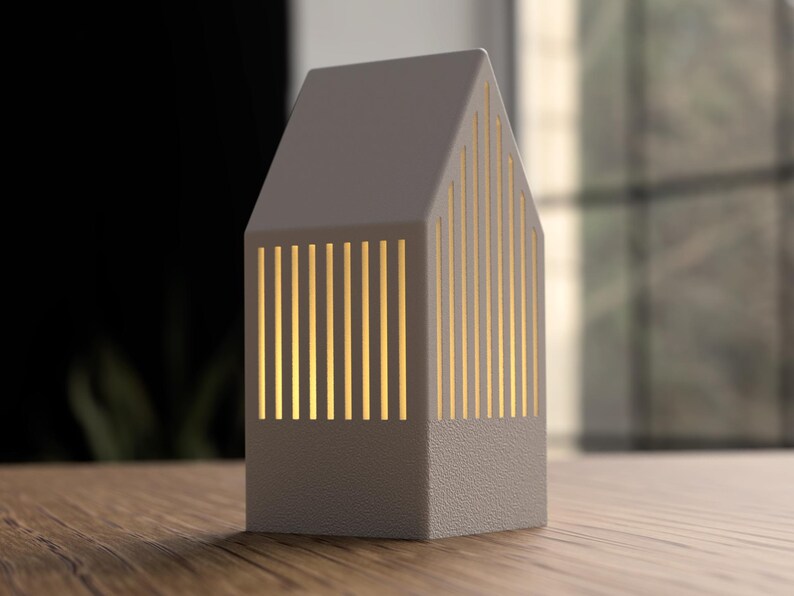 May include: A gray, house-shaped lamp with vertical, illuminated slots. The lamp has a textured base and a pitched roof. The warm light shines through the slots, creating a cozy ambiance. The lamp is on a wooden surface.