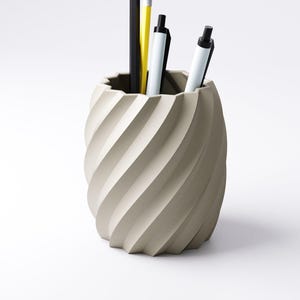 May include: A beige, spiral-shaped desk organizer holding a black pencil, a yellow pencil, and two white pens with black accents. The organizer has a textured, ridged design and sits on a white surface.