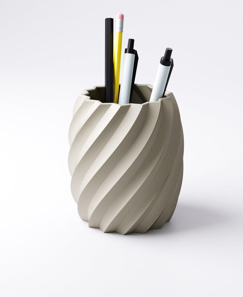 May include: A beige, spiral-shaped desk organizer holding a black pencil, a yellow pencil, and two white pens with black accents. The organizer has a textured, ridged design and sits on a white surface.