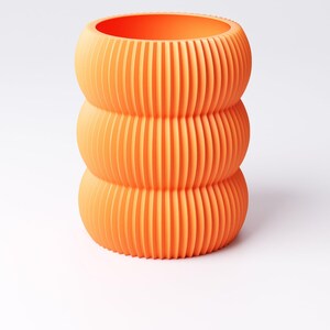May include: An orange, three-tiered vase with a ribbed design. The vase has a cylindrical shape with rounded sections, and the vertical ribs create a textured surface. The bright orange color gives it a modern look.
