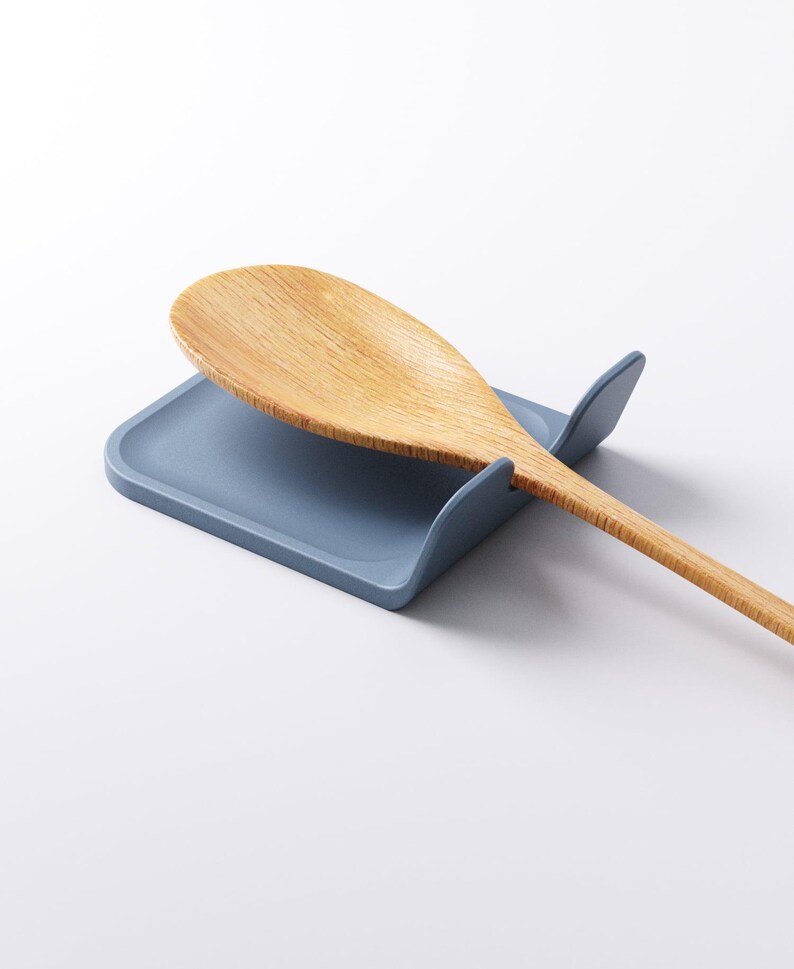 Op de afbeelding: Een lichtblauwe siliconen lepelhouder met een houten lepel. De lepelhouder heeft een rechthoekige basis met afgeronde randen en twee verhoogde zijkanten. De houten lepel is lichtbruin.