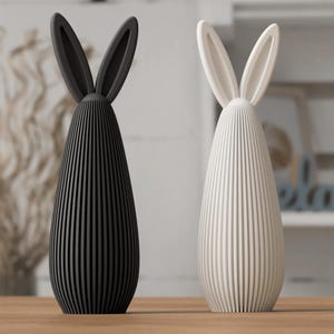 May include: Two decorative bunny figurines. One is black, the other is white. Each has long, upright ears and a vertically-lined body. They are placed on a wooden surface, suggesting a springtime or Easter theme.