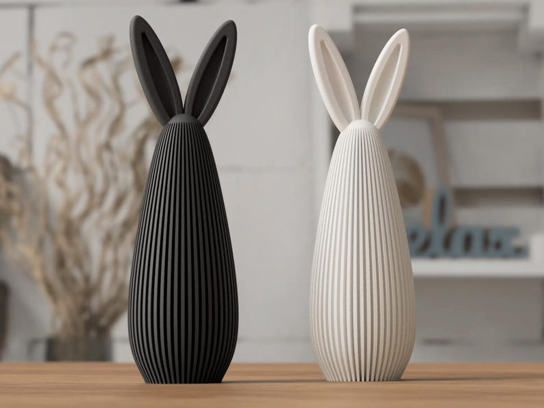 May include: Two decorative bunny figurines. One is black, the other is white. Each has long, upright ears and a vertically-lined body. They are placed on a wooden surface, suggesting a springtime or Easter theme.