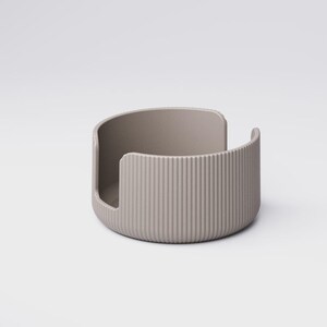 May include: A taupe-colored, semi-circular container with vertical ribbed detailing on the exterior. The open design features a curved back and a cutout on one side. The object is made of a solid material and sits on a white background.
