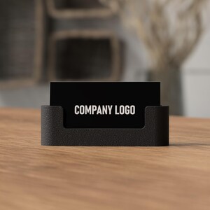 May include: A black business card holder with a textured finish, holding a black business card. The card displays the text "COMPANY LOGO" in white. The holder sits on a wooden surface.