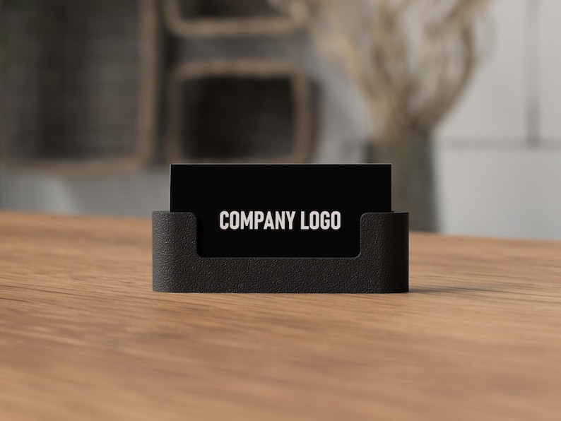 May include: A black business card holder with a textured finish, holding a black business card. The card displays the text "COMPANY LOGO" in white. The holder sits on a wooden surface.
