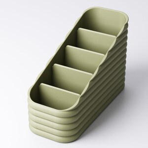 May include: A sage green desk organizer with four compartments. The organizer has a curved, ridged exterior and a stepped design. The compartments are rectangular and the overall shape is a rounded rectangle. The organizer is made of a solid, matte material.