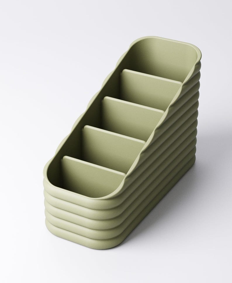 May include: A sage green desk organizer with four compartments. The organizer has a curved, ridged exterior and a stepped design. The compartments are rectangular and the overall shape is a rounded rectangle. The organizer is made of a solid, matte material.