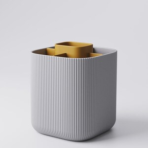 May include: A grey, ribbed, rectangular container with a mustard-yellow insert. The container has a rounded base and vertical lines. The insert has a square shape with rounded corners. It's designed for organizing items, likely for a desk.