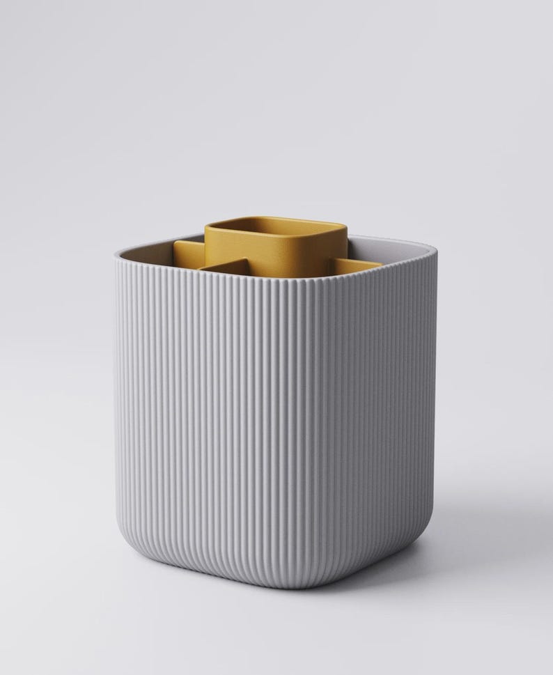 May include: A grey, ribbed, rectangular container with a mustard-yellow insert. The container has a rounded base and vertical lines. The insert has a square shape with rounded corners. It's designed for organizing items, likely for a desk.