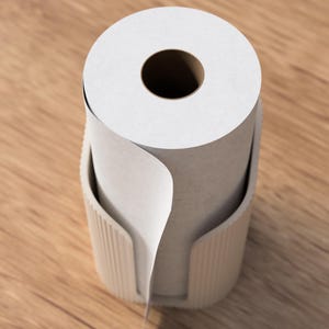 Op de afbeelding: Een rol witte papieren handdoeken zit in een beige houder. De papieren handdoekrol heeft een groot centraal gat. De houder heeft een gebogen ontwerp met verticale lijnen. De achtergrond is een lichtbruin houten oppervlak.