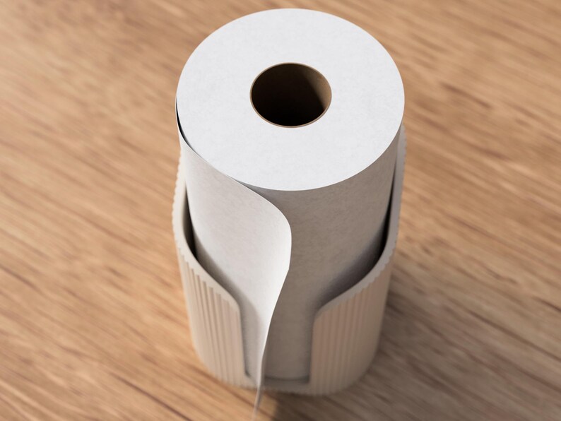 Op de afbeelding: Een rol witte papieren handdoeken zit in een beige houder. De papieren handdoekrol heeft een groot centraal gat. De houder heeft een gebogen ontwerp met verticale lijnen. De achtergrond is een lichtbruin houten oppervlak.