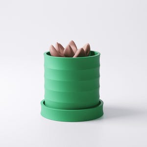 Op de afbeelding: Een groene keramische plantenpot met een geribbeld ontwerp, met een cluster van roze, bloembladachtige objecten. De pot staat op een bijpassende groene schotel. Het ontwerp is modern en minimalistisch.