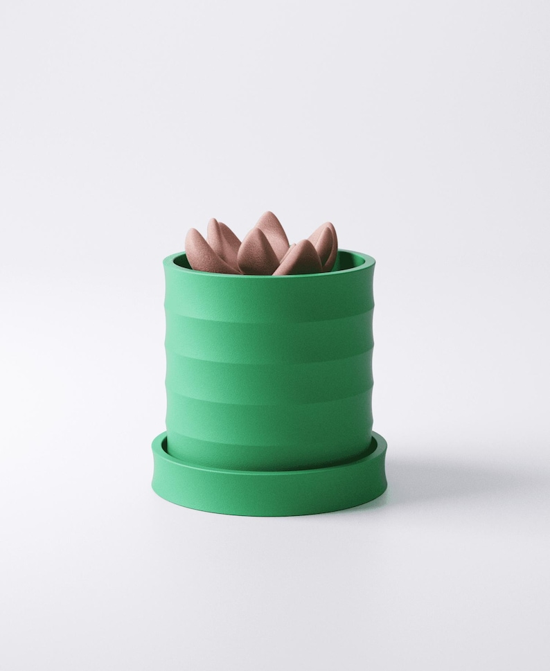 Op de afbeelding: Een groene keramische plantenpot met een geribbeld ontwerp, met een cluster van roze, bloembladachtige objecten. De pot staat op een bijpassende groene schotel. Het ontwerp is modern en minimalistisch.