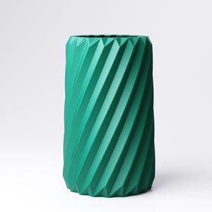 May include: A teal-colored, cylindrical vase with a geometric, ridged design. The vase has a textured surface with diagonal lines that create a spiral effect. The vase is a solid color and has a modern aesthetic.