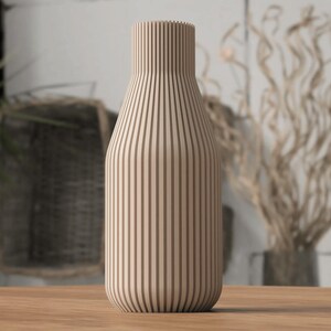 May include: A beige wooden vase with vertical brown stripes. The vase has a rounded base and a narrow neck. The vase is sitting on a wooden surface. The background includes a plant and decorative branches.