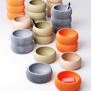 May include: A collection of modern, ribbed, cylindrical containers in various colors, including orange, beige, gray, and olive green. Some containers are stacked, and some hold pens or small objects. The containers have a minimalist design.