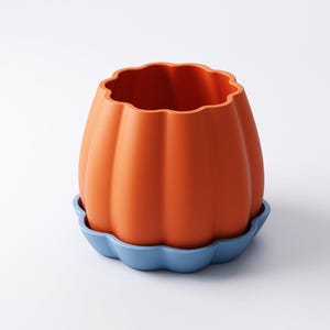 May include: An orange pumpkin-shaped planter with a wavy rim, sitting on a blue, scalloped saucer. The planter has vertical ridges and a smooth, matte finish. The design is simple and modern, suitable for indoor or outdoor use.
