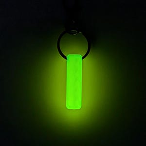 May include: A glowing neon green rectangular keychain pendant hangs from a black metal ring. The pendant has a textured surface and emits a bright, vibrant light against a dark background. The keychain is a simple, yet eye-catching accessory.
