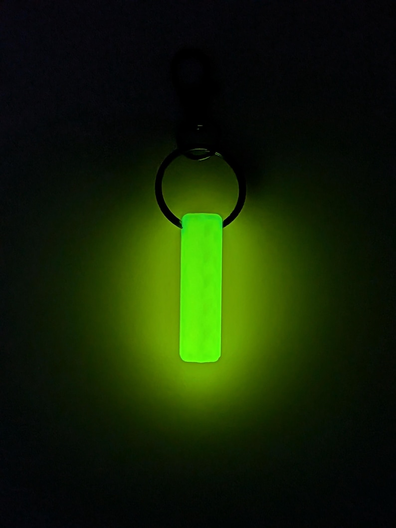 May include: A glowing neon green rectangular keychain pendant hangs from a black metal ring. The pendant has a textured surface and emits a bright, vibrant light against a dark background. The keychain is a simple, yet eye-catching accessory.