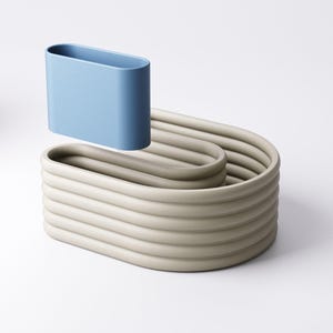 May include: A light blue, oval container hovers above a beige, wave-like, ribbed structure. The design has a minimalist aesthetic, with smooth, rounded forms and a neutral colour palette. The objects appear to be made of ceramic or a similar material.