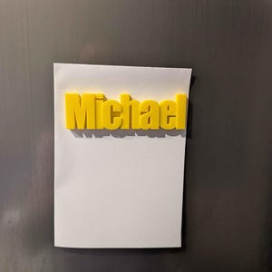 May include: A yellow 3D-printed name, "Michael," is affixed to a white piece of paper. The name is in a bold, sans-serif font. The paper is attached to a dark, metallic surface.