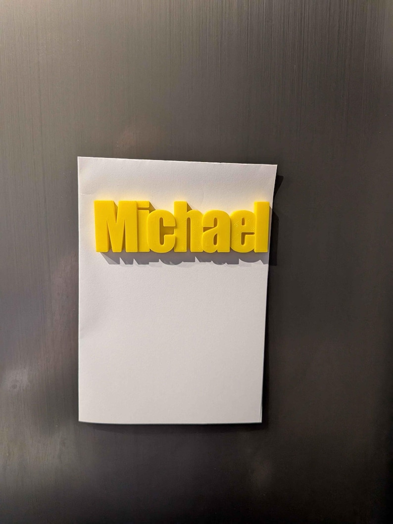 May include: A yellow 3D-printed name, "Michael," is affixed to a white piece of paper. The name is in a bold, sans-serif font. The paper is attached to a dark, metallic surface.
