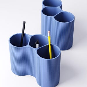 May include: Two blue desk organizers with a ribbed texture. Each organizer has three compartments of varying sizes. One organizer holds a black pen, a white pen, and a yellow pencil. The other organizer holds a black pen. The background is white.