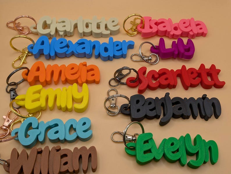 May include: A collection of colorful name keychains in various colors, including pink, blue, green, and brown. Each keychain features a different name in a bold, playful font, attached to a metal keyring.