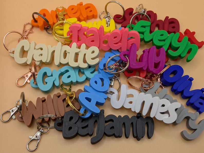 May include: A collection of colorful name keychains in various colors, including orange, pink, green, and blue. Each keychain features a different name in a bold, 3D font, attached to a metal keyring.