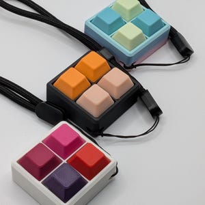 May include: Three square keycap pendants with black lanyards. One pendant has orange and peach keycaps, another has pink and red keycaps, and the last has blue and green keycaps. The pendants are on a white surface.
