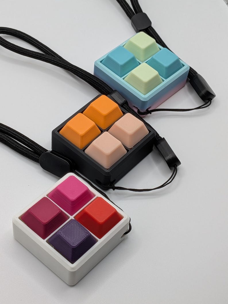 May include: Three square keycap pendants with black lanyards. One pendant has orange and peach keycaps, another has pink and red keycaps, and the last has blue and green keycaps. The pendants are on a white surface.