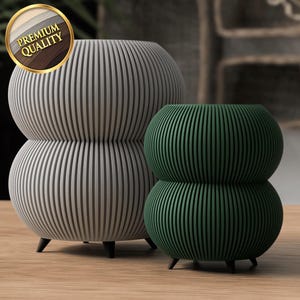 May include: Two ceramic planters with a ribbed design. The larger planter is grey and the smaller planter is green. Both planters have a rounded shape and three legs.
