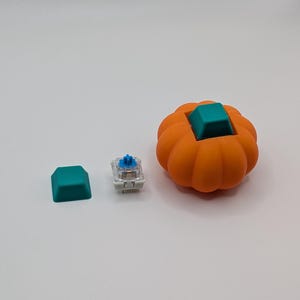 May include: An orange pumpkin-shaped keyboard keycap with a teal square keycap insert. A teal keycap and a mechanical keyboard switch are next to the pumpkin keycap. The items are on a white surface.