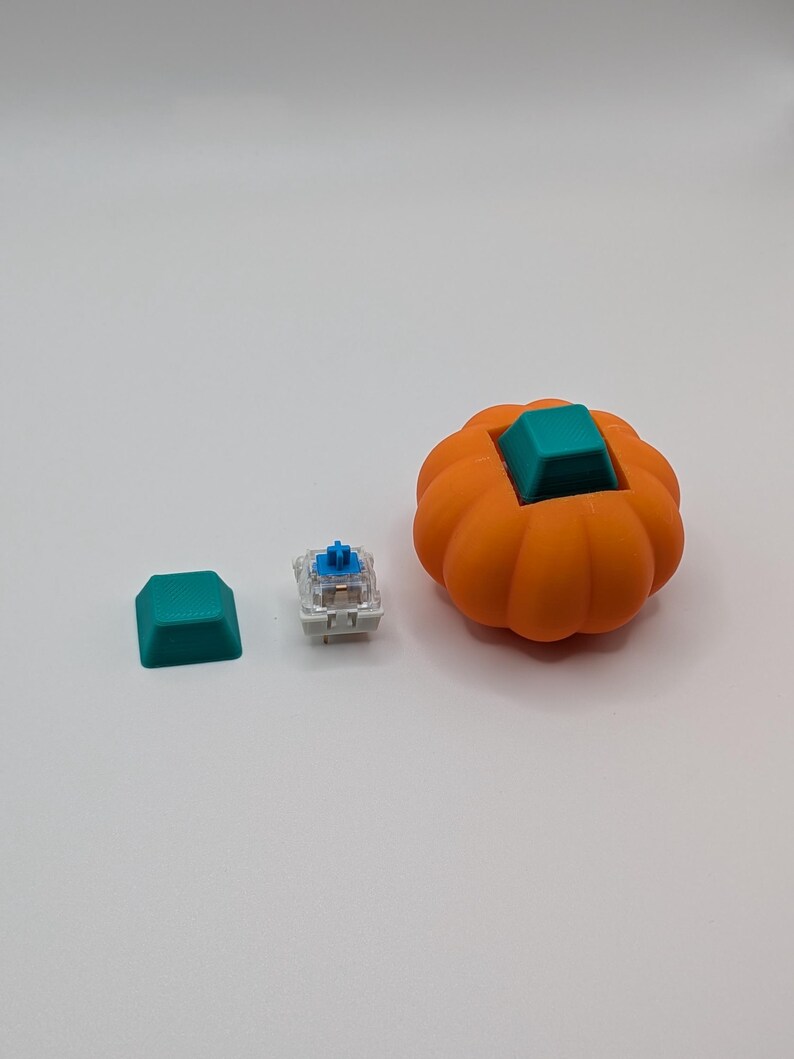 May include: An orange pumpkin-shaped keyboard keycap with a teal square keycap insert. A teal keycap and a mechanical keyboard switch are next to the pumpkin keycap. The items are on a white surface.