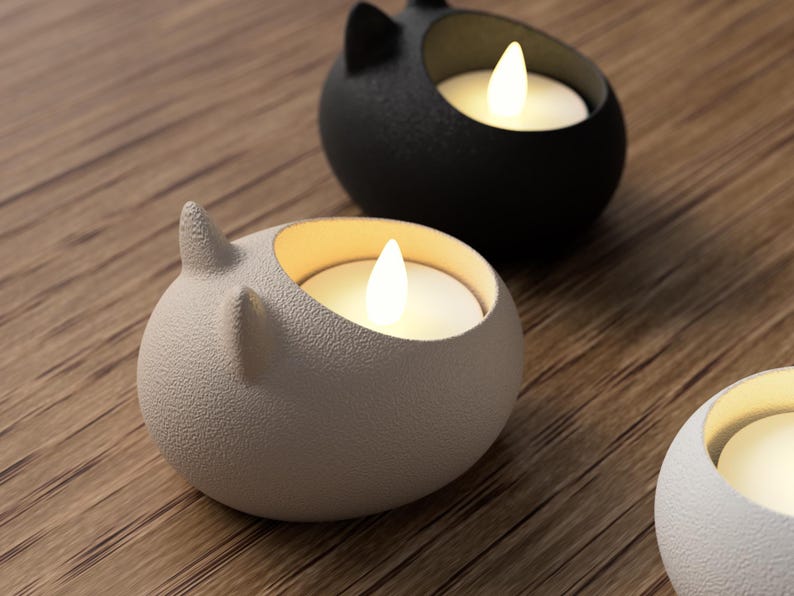 May include: A close-up of three cat-shaped candle holders in a warm, inviting setting. The candle holders are in shades of gray, black, and white, each with a glowing, warm-toned LED candle inside. The holders have a textured surface and are placed on a wooden surface.